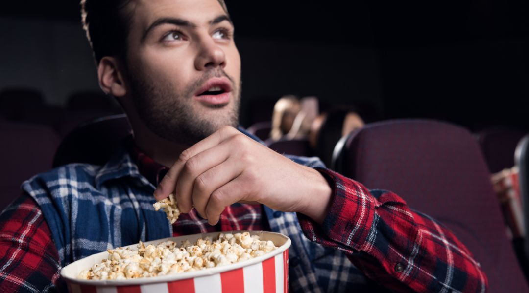 Homme assis dans une salle de cinéma tenant un seau de popcorn rayé, avec une expression de surprise ou de fascination