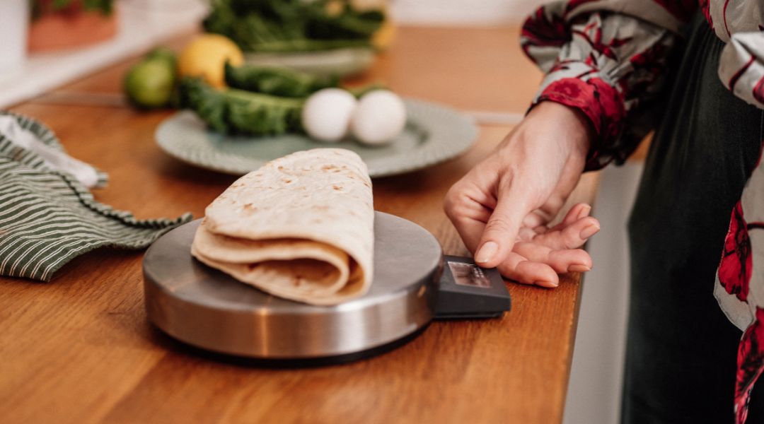 Gros plan d'une tortilla pesée sur une balance de cuisine, avec des légumes frais et des œufs en arrière-plan