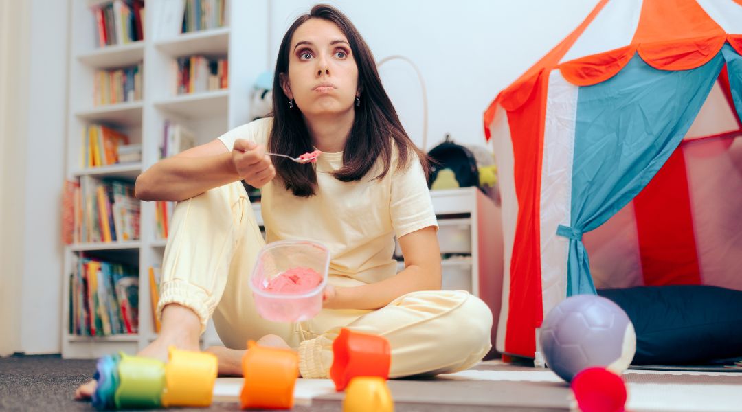 Femme assise par terre dans une salle de jeux, mangeant de la glace directement dans un pot, entourée de jouets