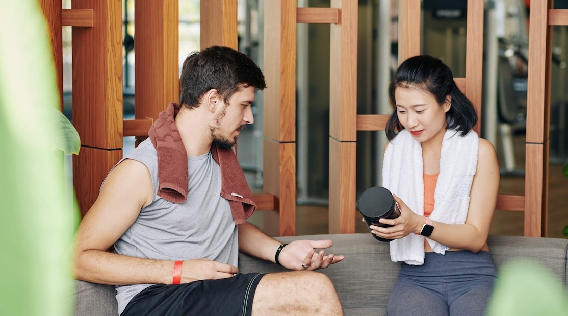 Un homme et une femme en tenue de sport assis sur un banc, discutant d'un contenant de supplément qu'elle tient, dans une salle de sport.