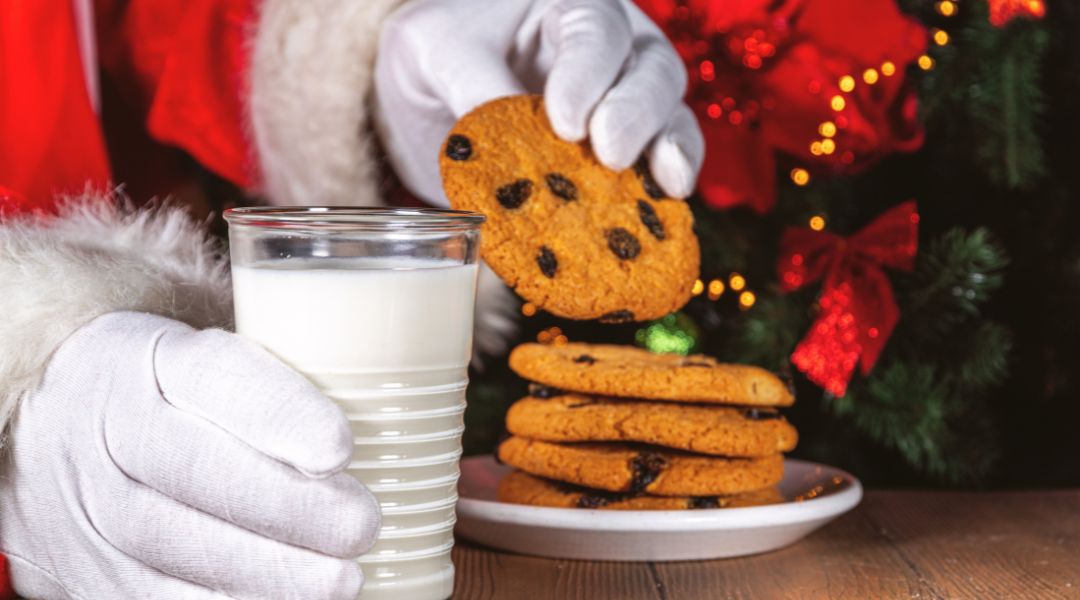 La main gantée du Père Noël tenant un biscuit au-dessus d’un verre de lait, avec une pile de biscuits et des décorations de Noël en arrière-plan.