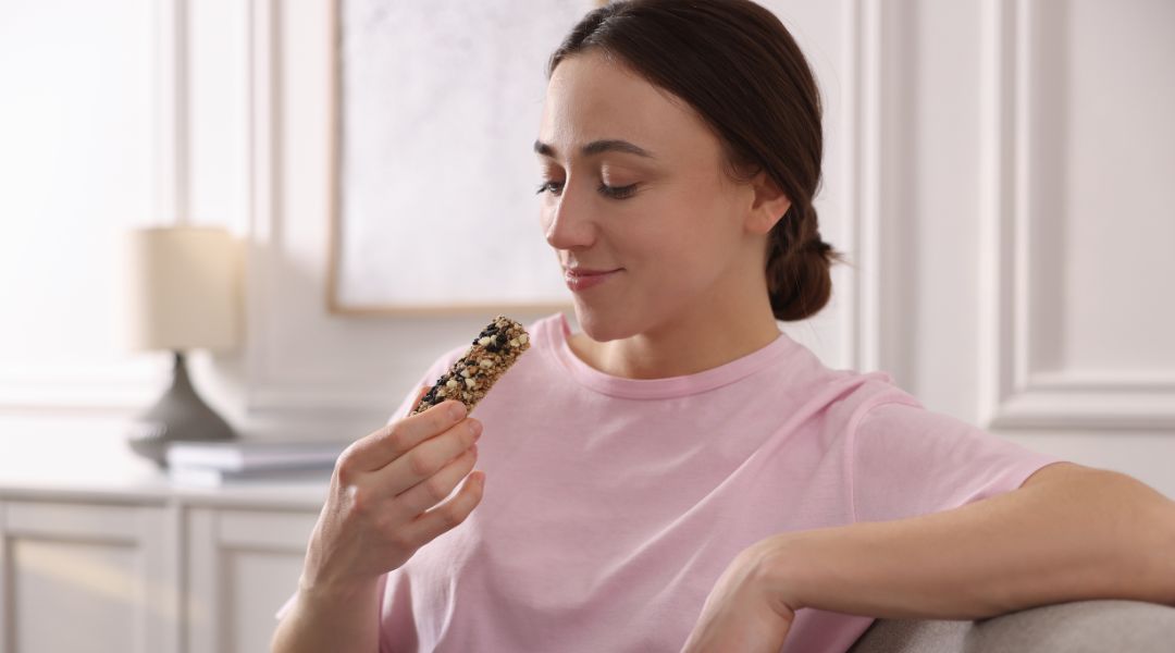 Femme en t-shirt rose assise sur un canapé, souriant en tenant une barre de céréales