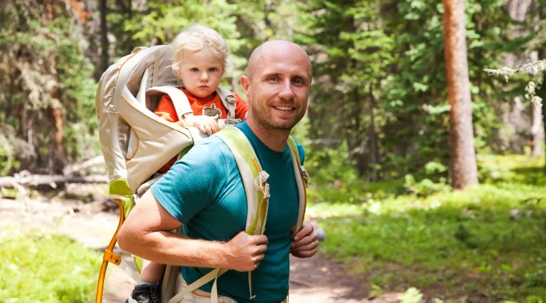 Père souriant portant son enfant dans un sac de randonnée en forêt