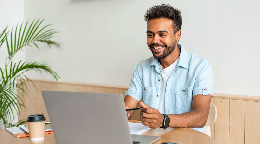 Homme souriant devant son ordinateur portable, travaillant avec une tasse de café et un carnet sur le bureau dans un cadre confortable