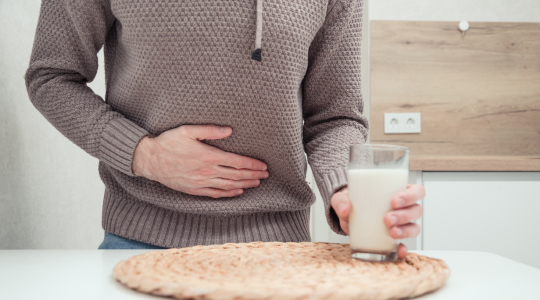 Personne tenant son ventre avec une main, exprimant un inconfort, tout en tenant un verre de lait de l'autre main, posée sur une table.