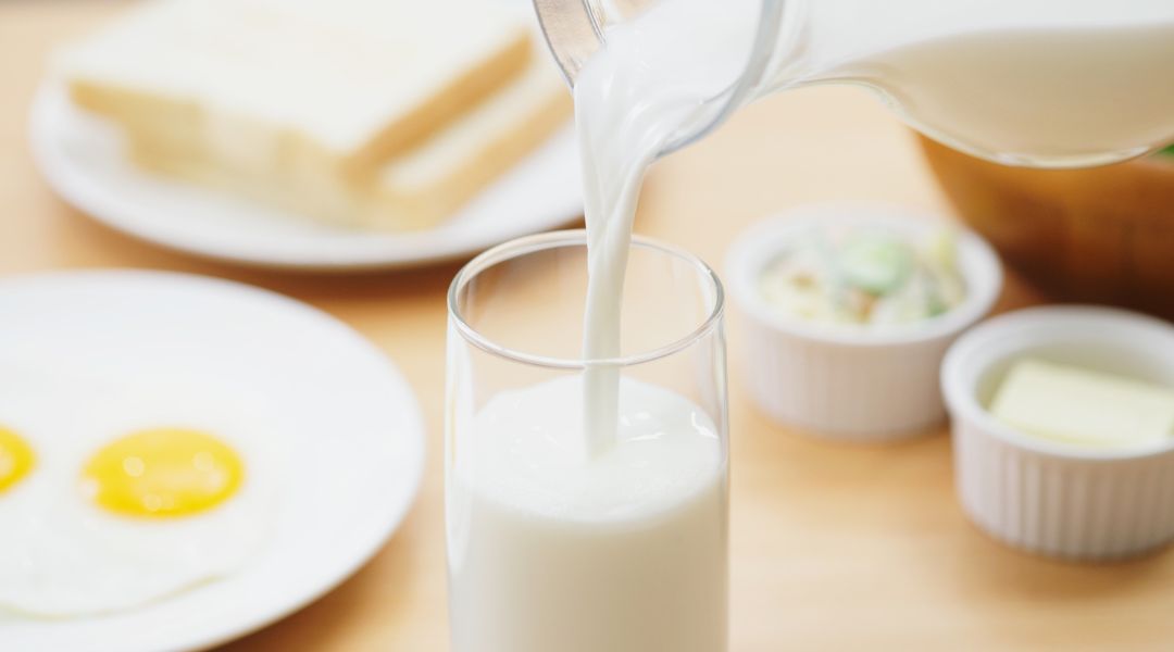 Milk being poured from a container into a clear glass on a wooden table, with a fried egg on a plate and small bowls of condiments nearby.