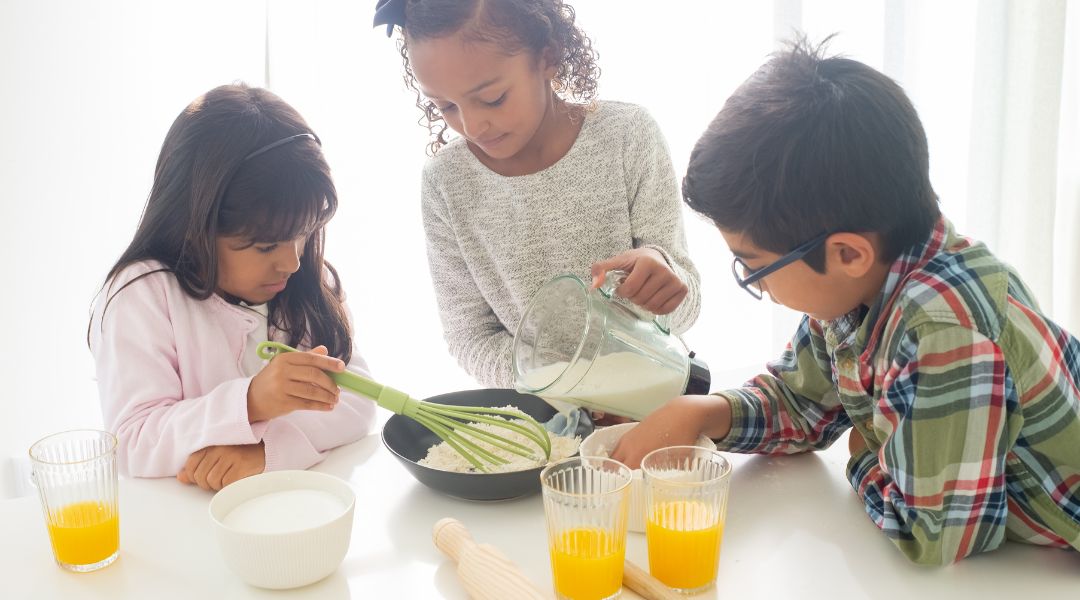 Three kids in a bright kitchen cracking eggs and whisking ingredients together in a bowl, with cups of orange egg mixture and a wooden rolling pin on the table.