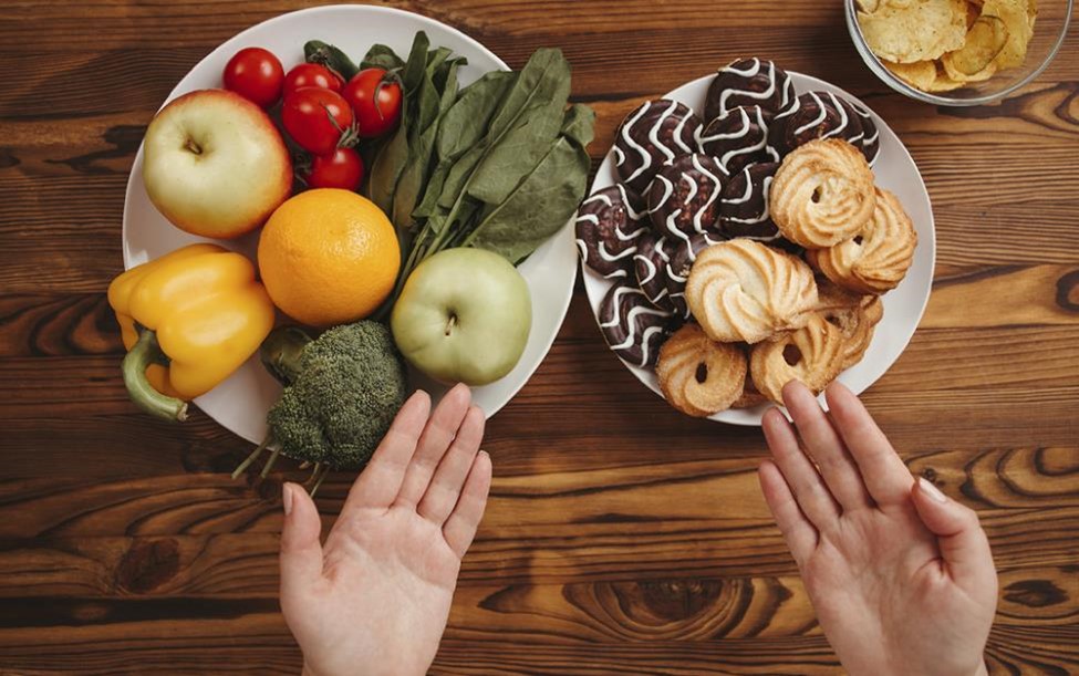 Deux assiettes sur une table en bois, l'une avec des fruits et légumes, l'autre avec des biscuits et pâtisseries, avec des mains désignant les deux.  Two plates on a wooden table, one with fruits and vegetables, the other with cookies and pastries, with hands gesturing towards both.