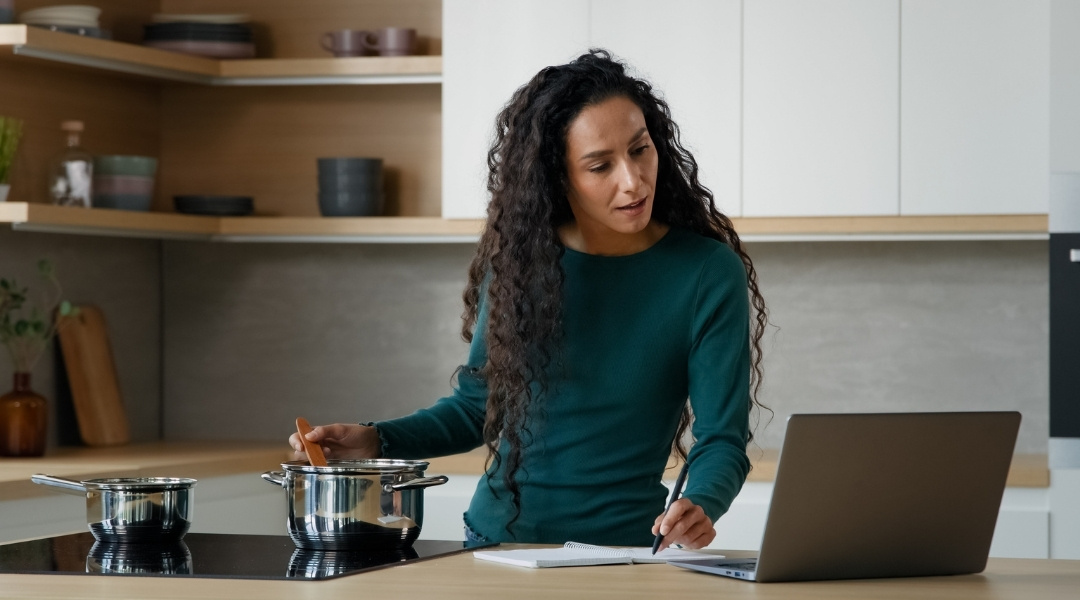 Une femme aux cheveux bouclés cuisine sur une plaque de cuisson moderne tout en regardant un ordinateur portable et en prenant des notes dans un carnet.