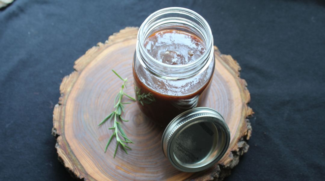 Pot en verre de sauce barbecue foncée avec une branche de romarin sur une surface en bois rustique, couvercle à côté