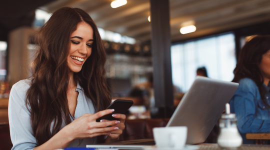 Jeune femme souriante assise dans un café, utilisant un téléphone portable tout en ayant un ordinateur portable ouvert devant elle.