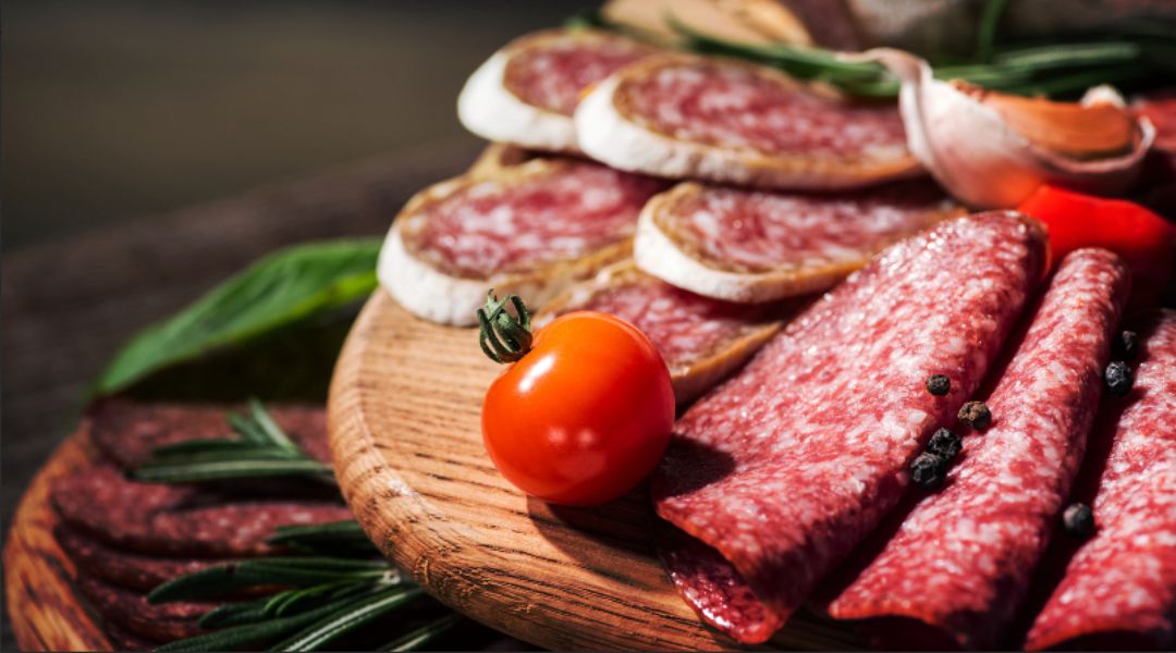 Plateau en bois avec du salami tranché, des viandes séchées et du camembert, accompagnés de tomates cerises, de branches de romarin et de grains de poivre.