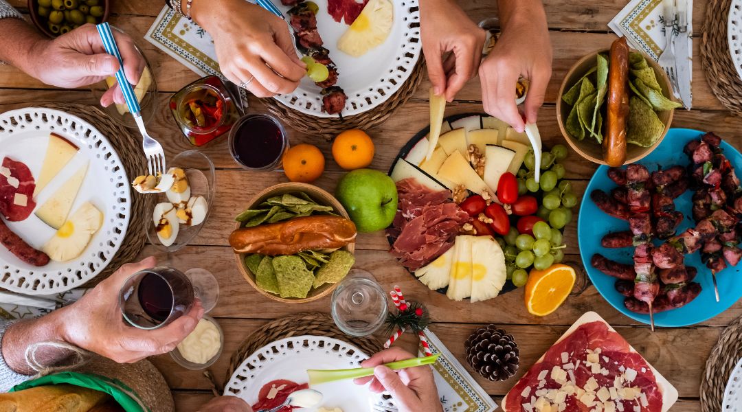 Table festive garnie d'assiettes de fromages, charcuteries, brochettes grillées, fruits frais, noix et sauces, entourée de convives se servant.