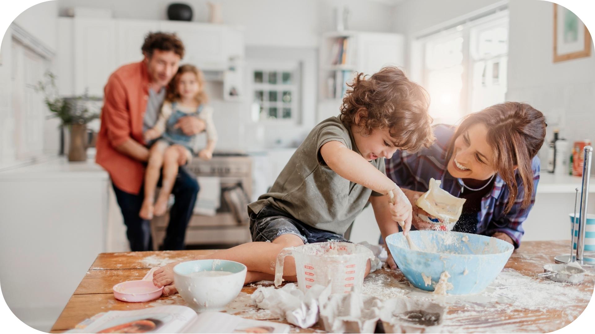 Une famille joyeuse cuisinant ensemble dans une cuisine lumineuse. Une mère et un jeune garçon mélangent des ingrédients dans un bol sur une table en désordre, tandis qu’un père tient une petite fille en arrière-plan