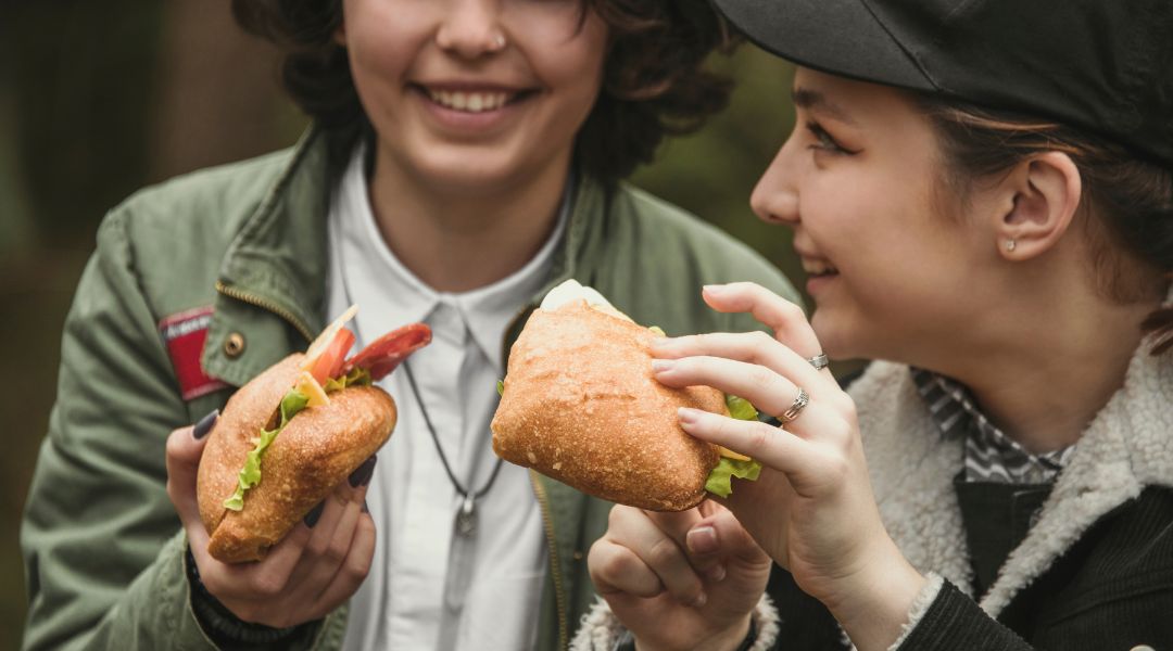 Deux jeunes femmes souriantes tenant des sandwichs garnis de laitue, de tomates et d'autres ingrédients frais, partageant un repas en plein air.