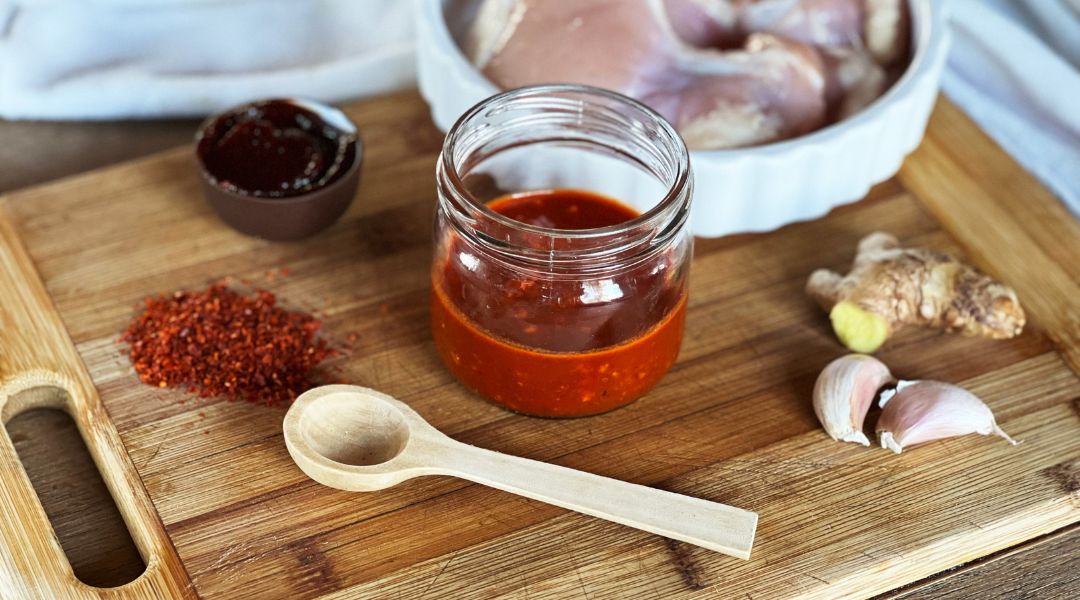 Pot de sauce rouge épicée sur une planche avec poudre de chili, gingembre frais, gousses d'ail et poulet cru en arrière-plan