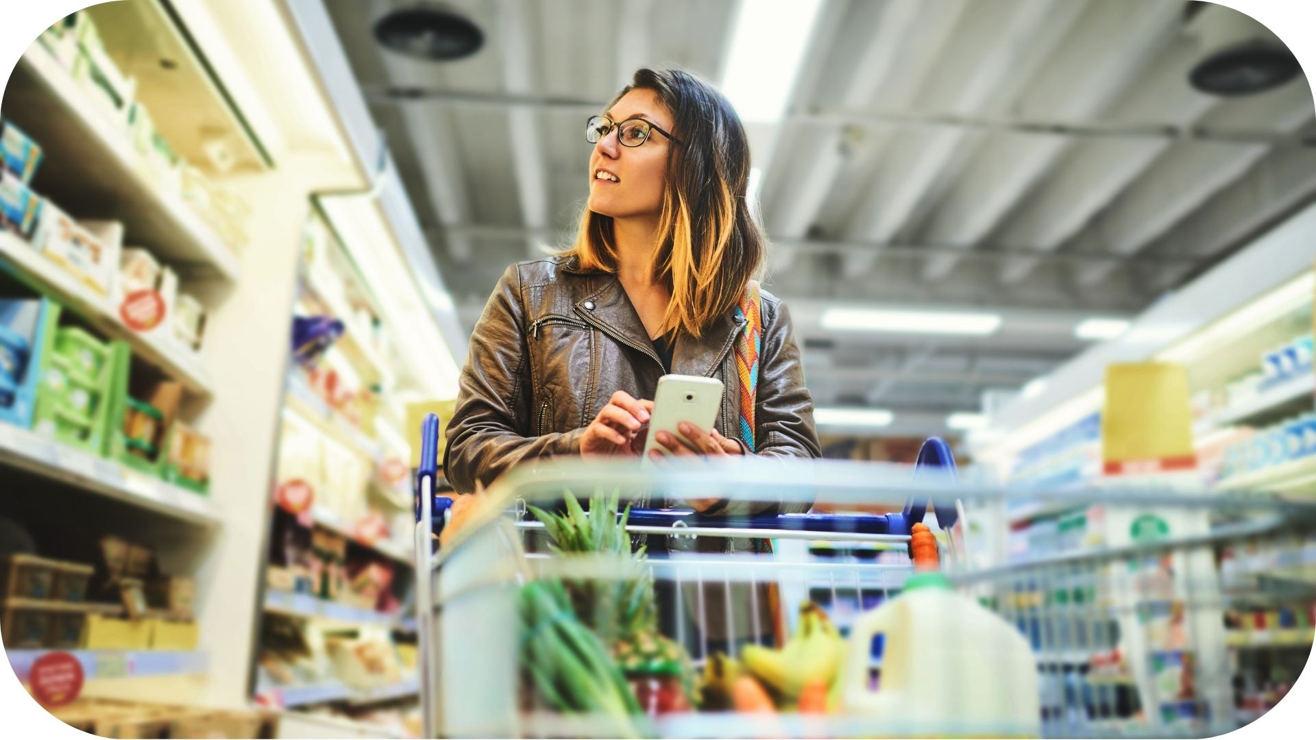 Femme faisant ses courses dans un supermarché, poussant un chariot avec des produits frais et tenant un smartphone