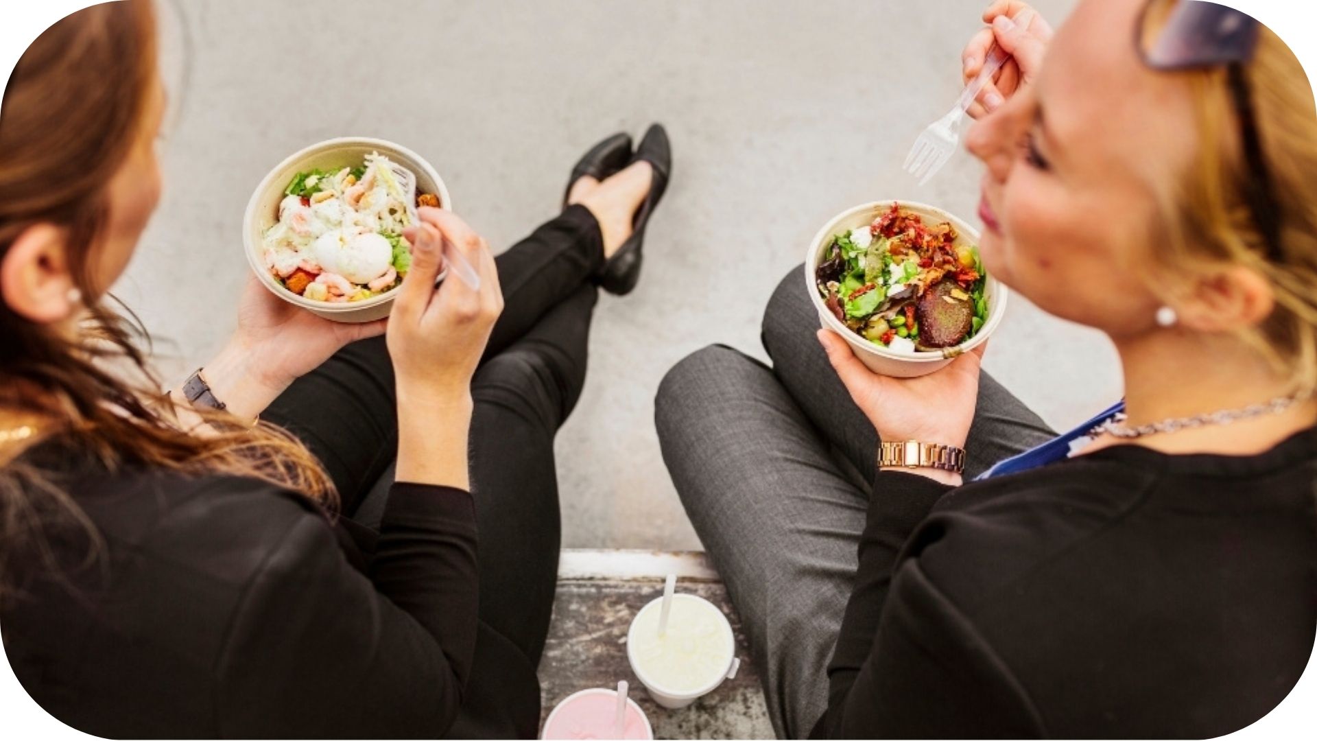 Deux femmes assises en train de manger des salades en plein air, chacune tenant une fourchette et discutant