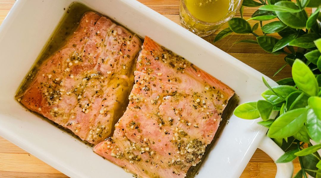  Deux filets de saumon marinés dans un plat blanc, garnis d'herbes, avec un petit verre de marinade à côté