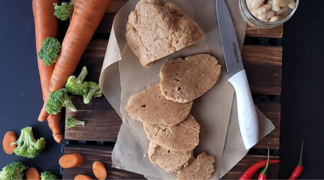 Viande végétalienne tranchée sur du papier sulfurisé, entourée de carottes fraîches, de brocolis, d'amandes et d'un couteau sur une surface en bois sombre.