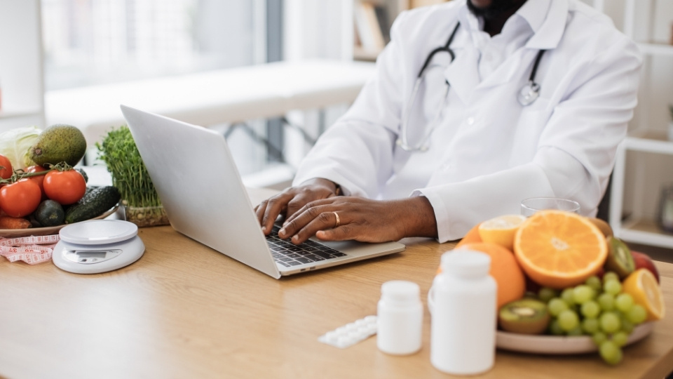 Un nutritionniste diététiste en blouse blanche travaille sur un ordinateur portable avec des fruits, légumes et suppléments sur la table.