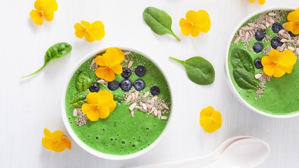 Un bol de smoothie vert vif garni de fleurs comestibles jaunes, de myrtilles fraîches, de graines de chia, de graines de tournesol et de feuilles de jeunes épinards. Le bol est disposé sur un fond clair avec des feuilles d'épinards et des fleurs jaunes supplémentaires éparpillées en décoration.