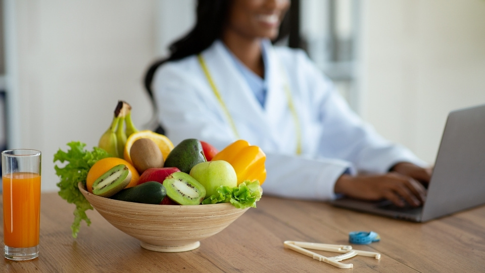 Un bol de fruits frais, dont kiwi, pomme et banane, avec un verre de jus sur une table, tandis qu'une nutritionniste travaille sur un ordinateur portable en arrière-plan.