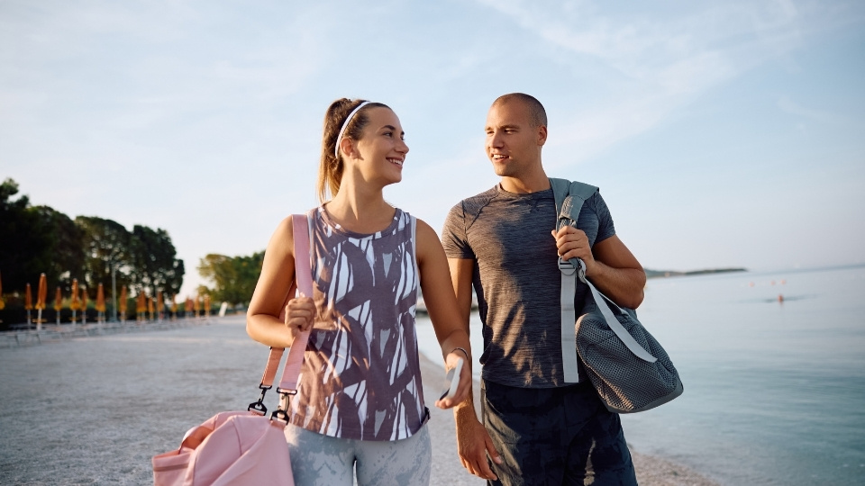 Un couple souriant en tenue de sport marche sur une plage, chacun portant un sac de sport, avec la mer et des arbres en arrière-plan.