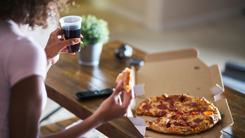 Personne tenant une part de pizza au pepperoni et un verre de soda, avec une boîte à pizza sur une table en bois.