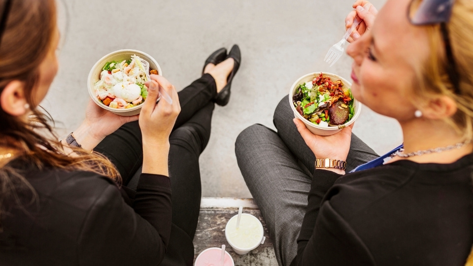 Deux femmes assises à l'extérieur, mangeant des bols de salade fraîche avec des légumes verts et garnitures, accompagnées de boissons.