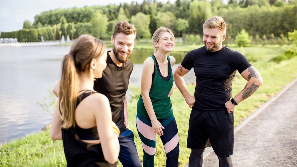 Un groupe de quatre amis en tenue de sport souriant et discutant à l'extérieur près d'un lac, avec une zone boisée en arrière-plan.