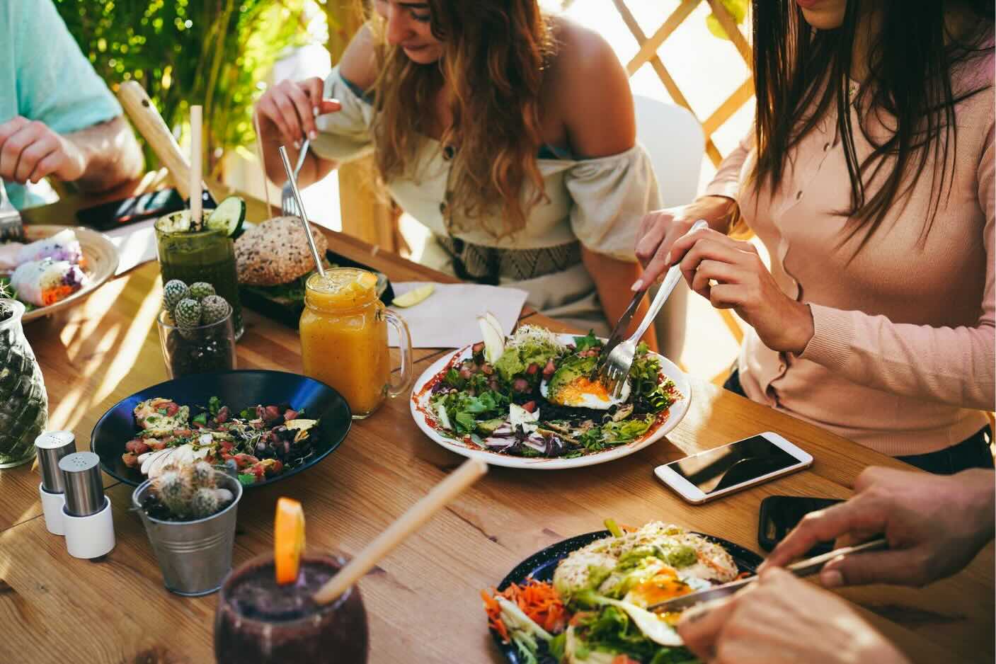 Group of people enjoying a meal with fresh salads, smoothies, and bread at an outdoor table.