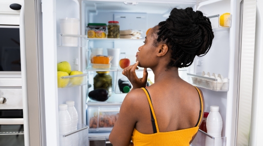 Femme debout devant un réfrigérateur ouvert, réfléchissant en regardant différents aliments et boissons à l'intérieur.  Woman standing in front of an open fridge, thoughtfully looking inside at various food items and drinks.