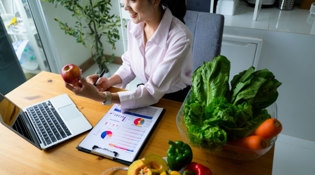 A person sitting at a table with a laptop, charts, a measuring tape, and a variety of fresh fruits and vegetables, including lettuce, carrots, bell peppers, and apples.