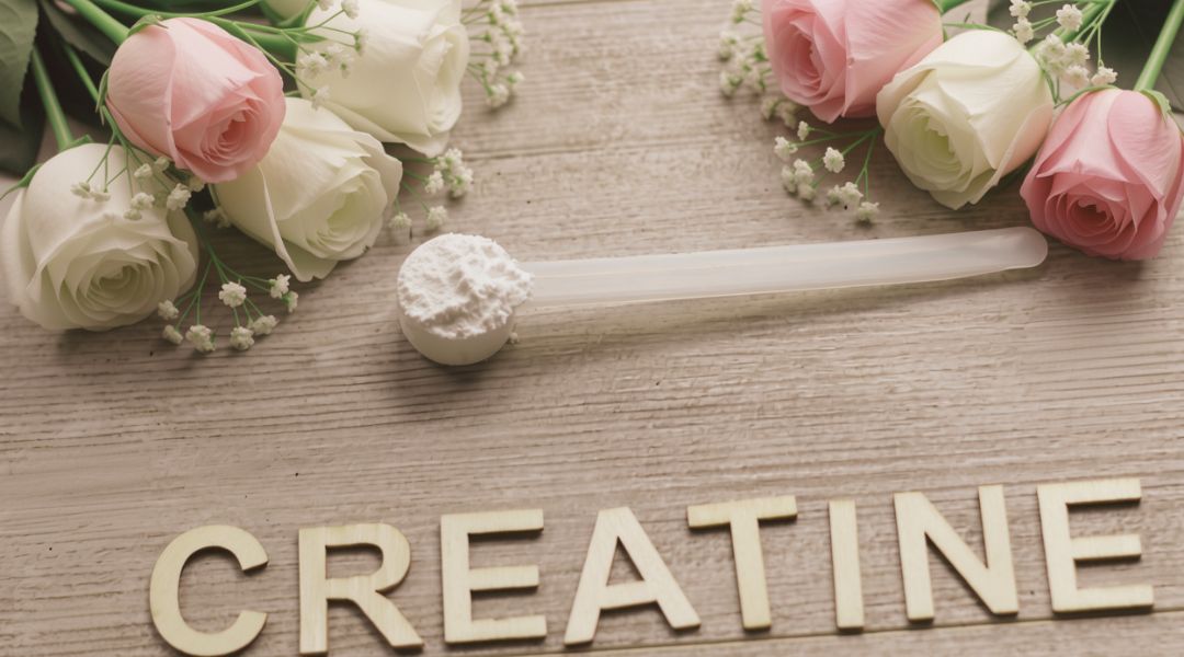 Cream and pink roses with small white blossoms arranged on a light wooden background next to a wooden spoon-shaped scoop and the word “CREATINE” in gold letters.