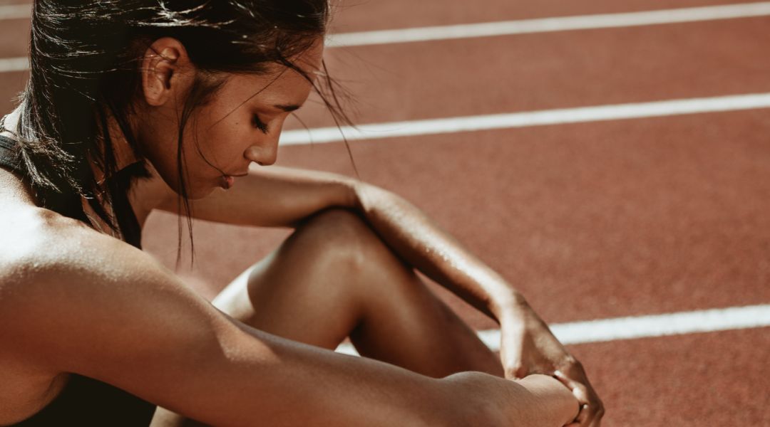A runner in a black sports top and shorts crouches on a track, fastening her shoelace while leaning forward, with white lane lines in the background.