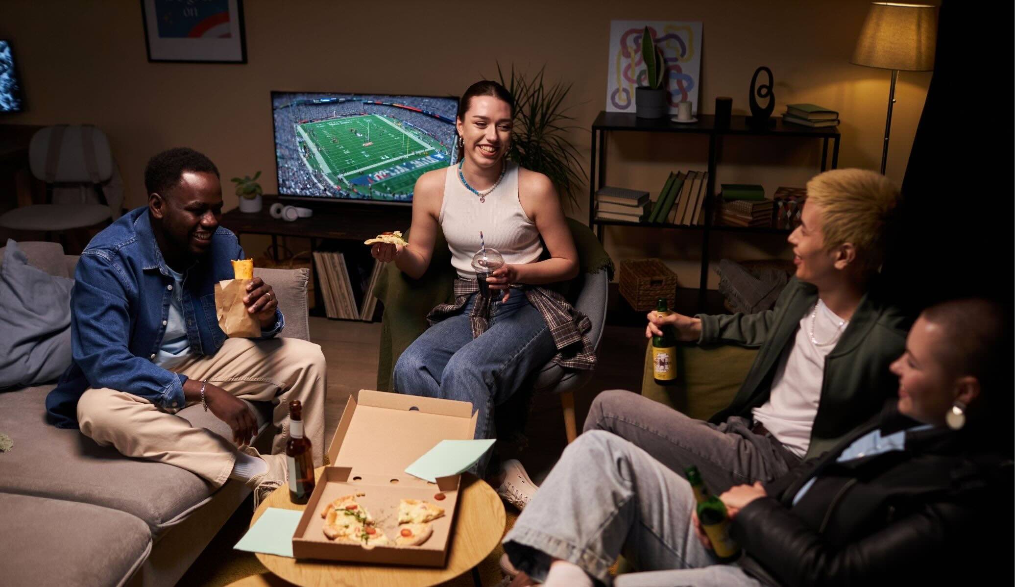 Quatre amis rient et mangent de la pizza dans un salon confortable en regardant un match de football américain à la télévision