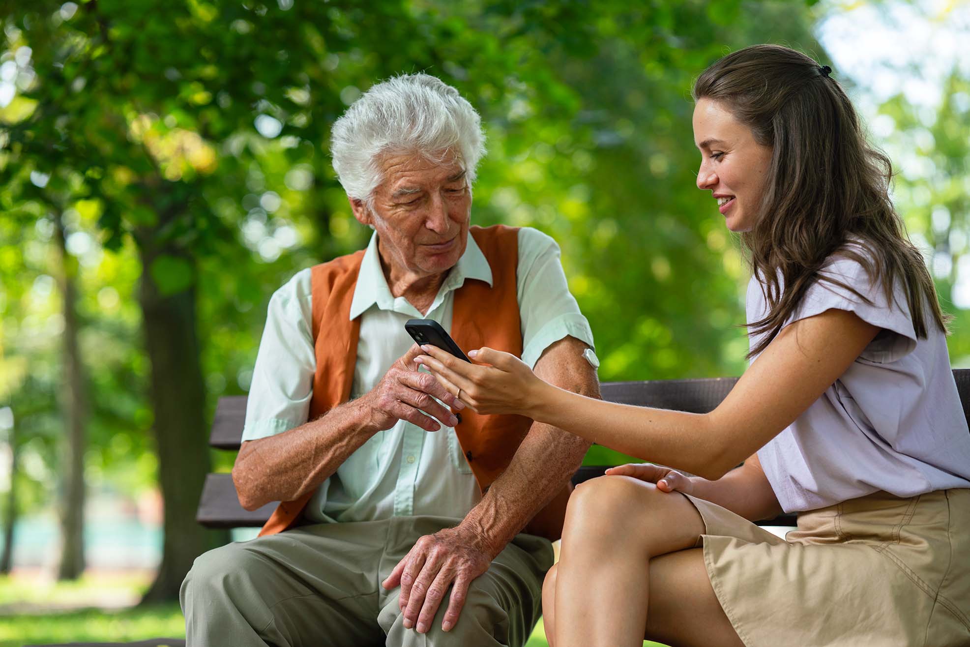 A young woman showing an elderly man how to use a smartphone while they sit on a park bench. The man has a continuous glucose monitor on his arm.