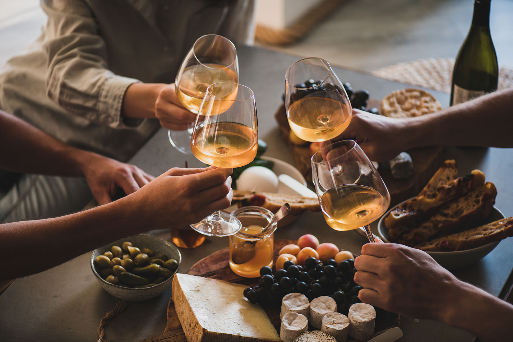 Friends toasting with white wine over a table of cheese, bread, grapes, and olives.