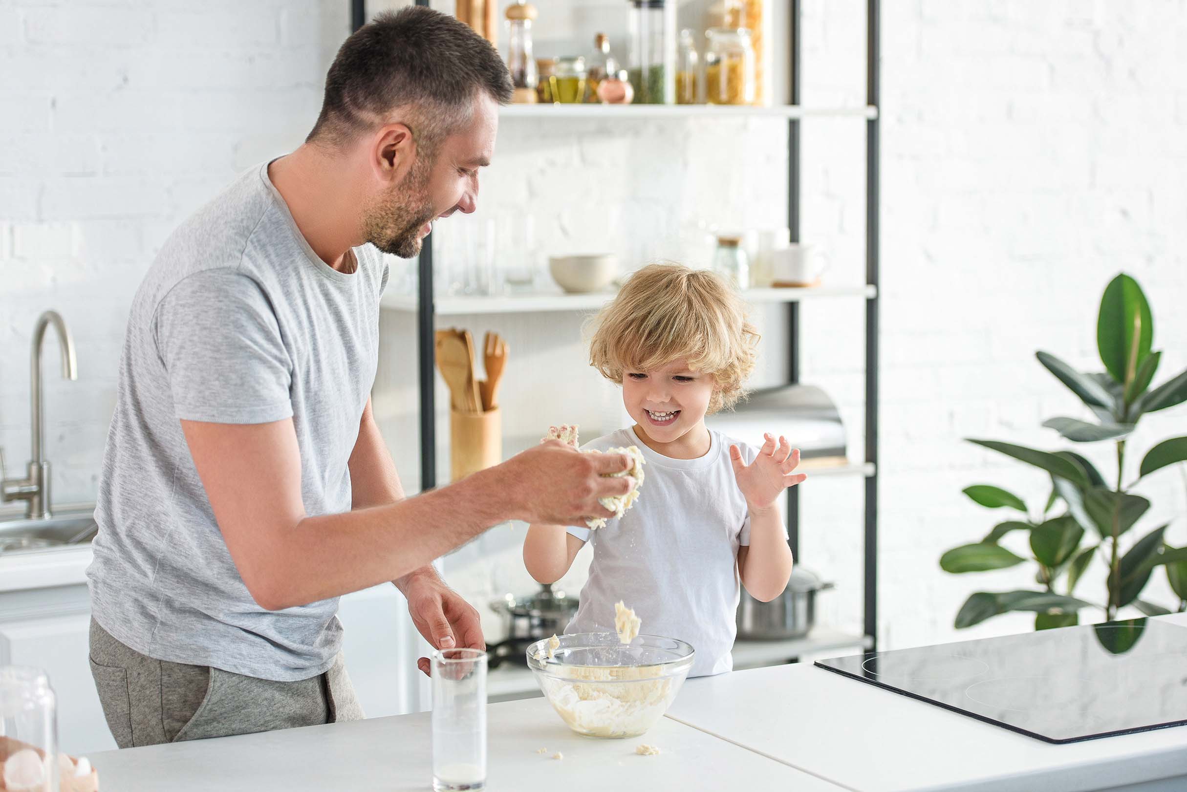 Un père et son fils rient et s'amusent en préparant de la pâte dans une cuisine moderne et lumineuse - A father and his son are laughing and having fun while making dough in a bright, modern kitchen