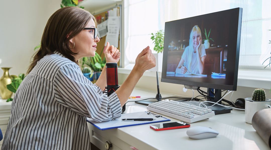 Une femme portant des lunettes gesticule pendant un appel vidéo avec une autre personne sur l’écran de son ordinateur, dans un bureau à domicile