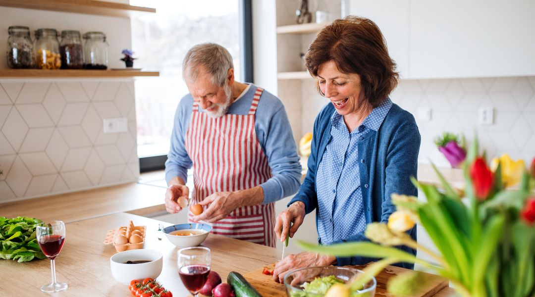 Un couple âgé souriant prépare des légumes et des œufs dans une cuisine lumineuse avec des tulipes sur le comptoir