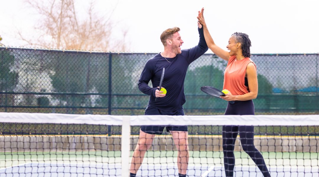 Deux personnes se tapent dans la main sur un court de pickleball, tenant des raquettes et des balles, célébrant une partie