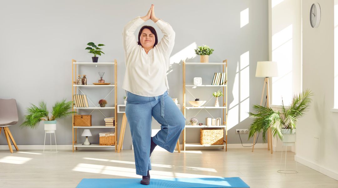 Une femme pratique le yoga dans un salon lumineux, en équilibre dans la posture de l’arbre sur un tapis bleu