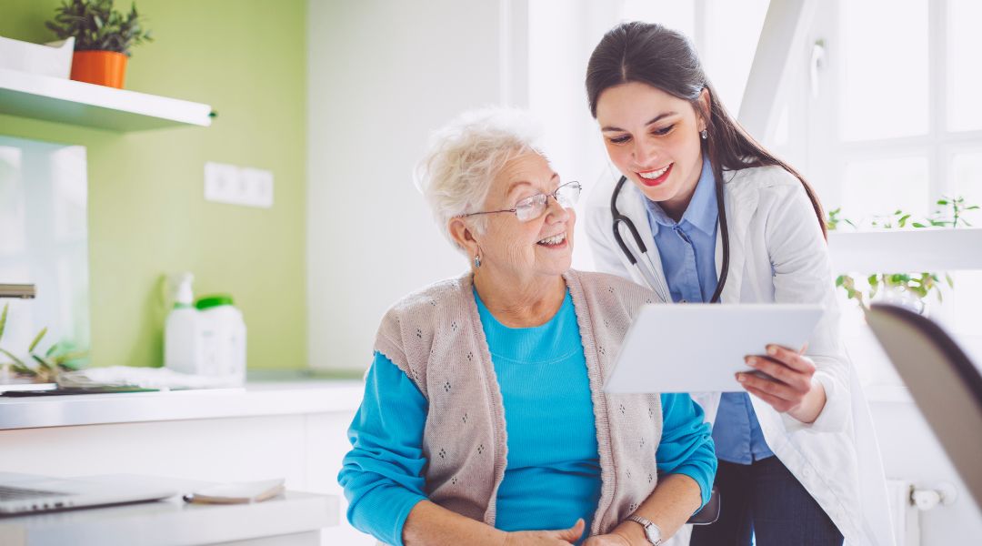 Une docteure montre une tablette numérique à une femme âgée, souriant ensemble dans un bureau moderne et lumineux