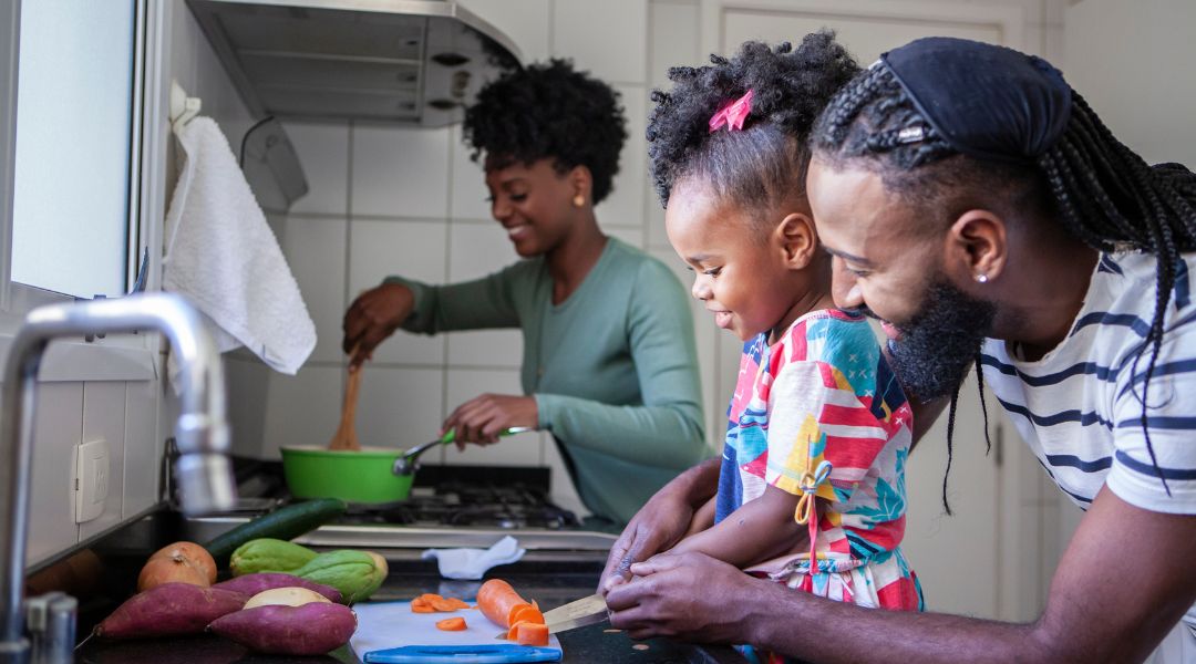 Une famille de trois personnes cuisine ensemble dans une cuisine, le père et la fille coupant des légumes tandis que la mère remue une casserole