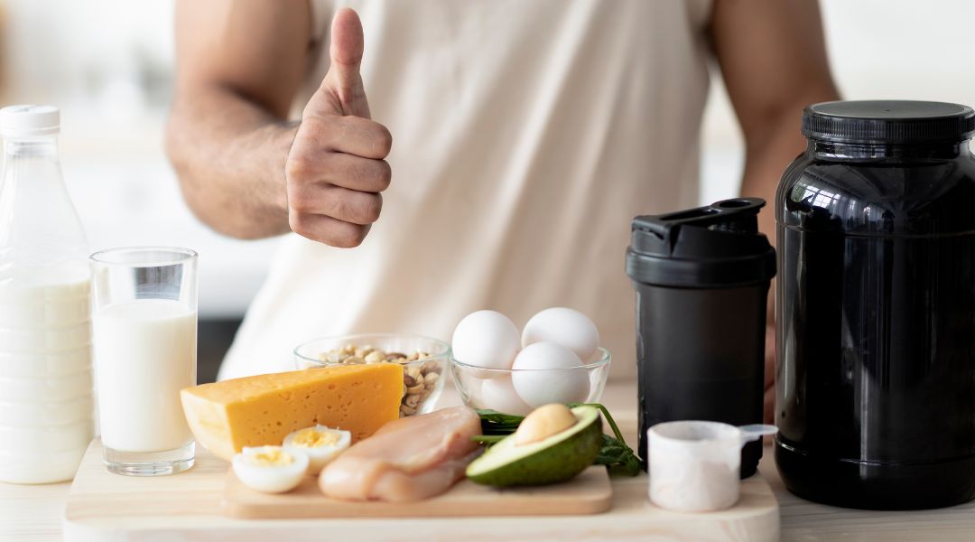 Homme faisant un pouce levé à côté d'une table avec des aliments riches en protéines, dont œufs, fromage, poulet et shake protéiné