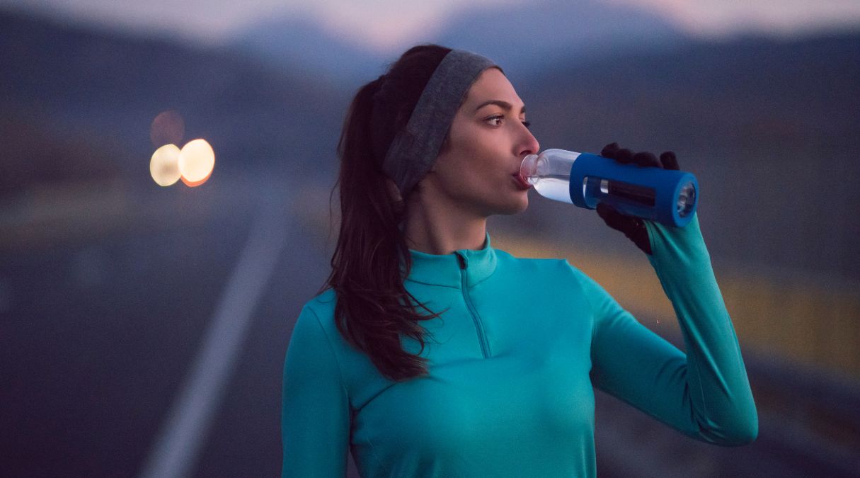 Femme buvant d'une bouteille en faisant de l'exercice en extérieur au crépuscule, portant une veste turquoise