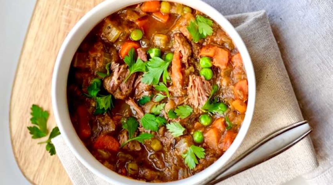 Un bol de ragoût de bœuf avec carottes, pois et persil frais sur une surface en bois - Recette ÉquipeNutrition