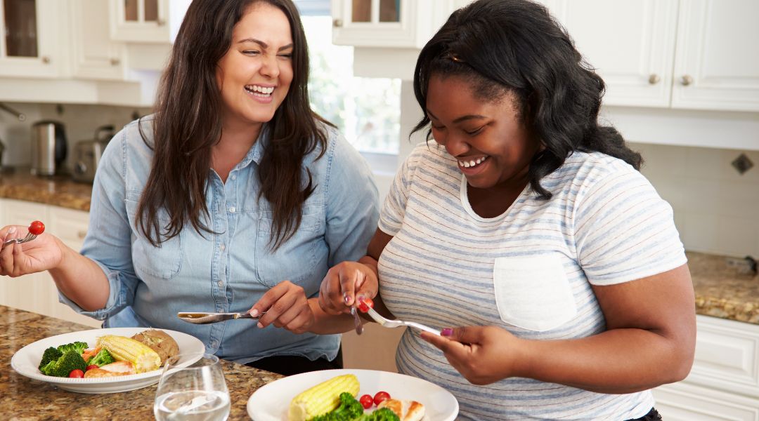 Deux femmes rient en mangeant un repas sain dans une cuisine, profitant de leur compagnie